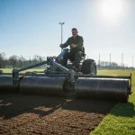 Pracownik przygotowujący podłoże na boisku sportowym do założenia trawnika - wyrównanie i walcowanie gruntu